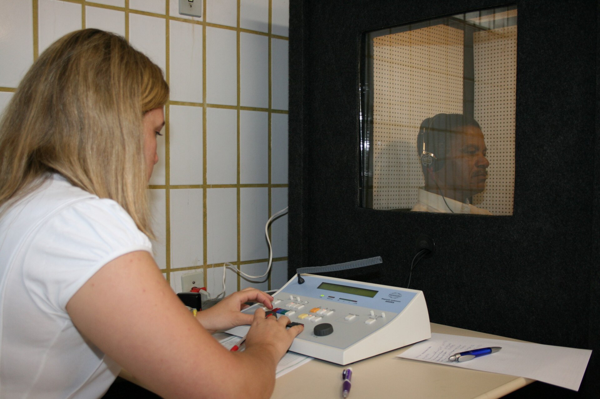 Profissional realiza exame auditivo com audiômetro e cabine acústica.